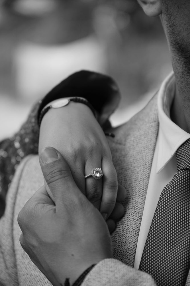 Black-and-white close-up of a couple holding hands, a round halo ring on her finger