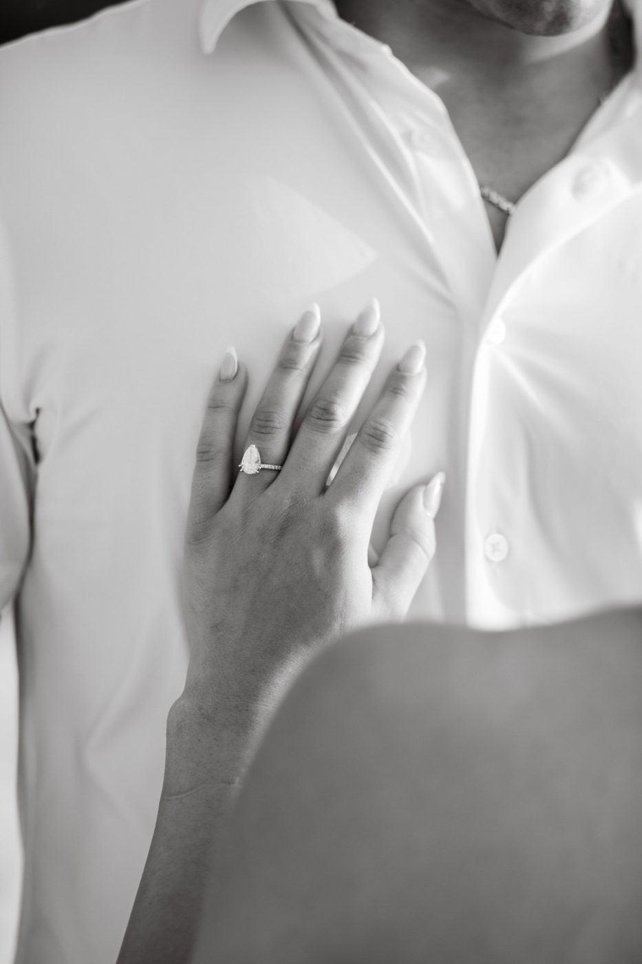 Black-and-white photo of a hand resting on a partner’s chest, wearing a pear-shaped diamond ring with a pavé band