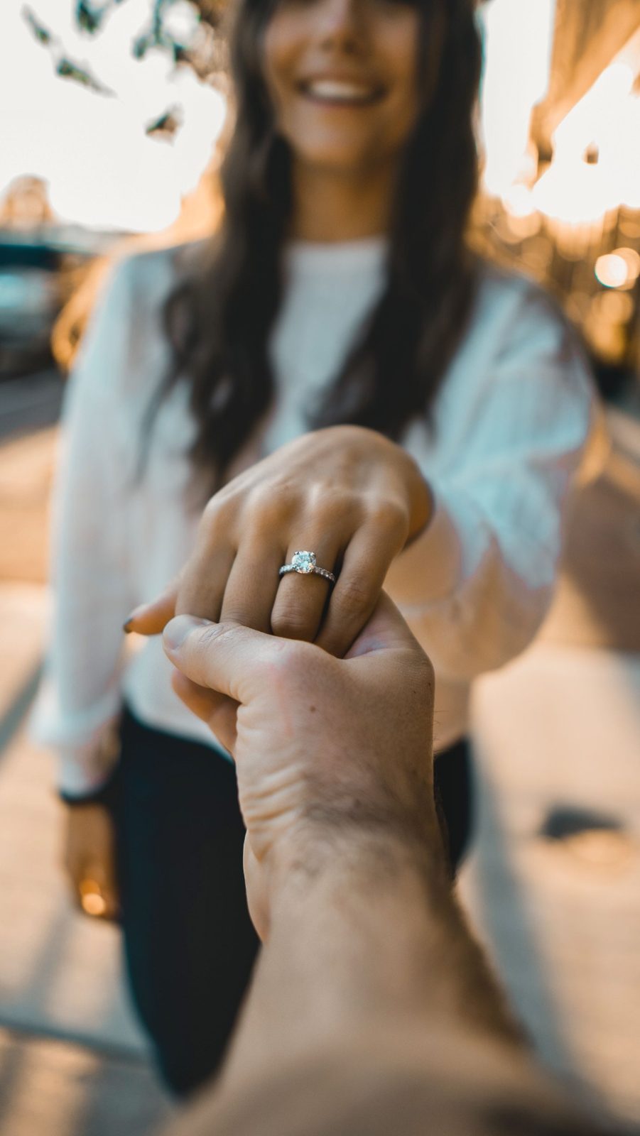 A smiling woman holds out her hand, showing a round diamond engagement ring