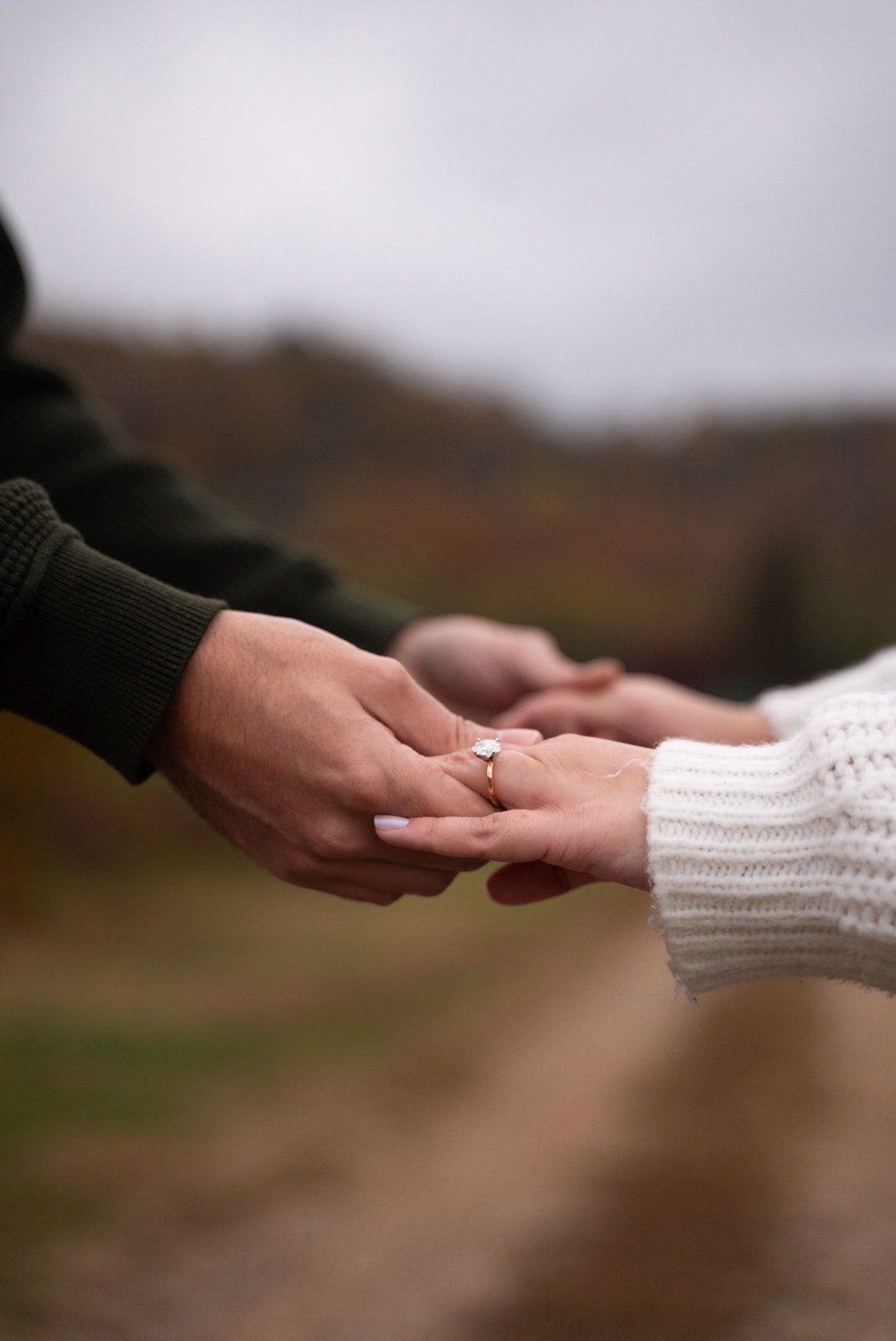 A couple holds hands outdoors in autumn, an oval solitaire on her finger