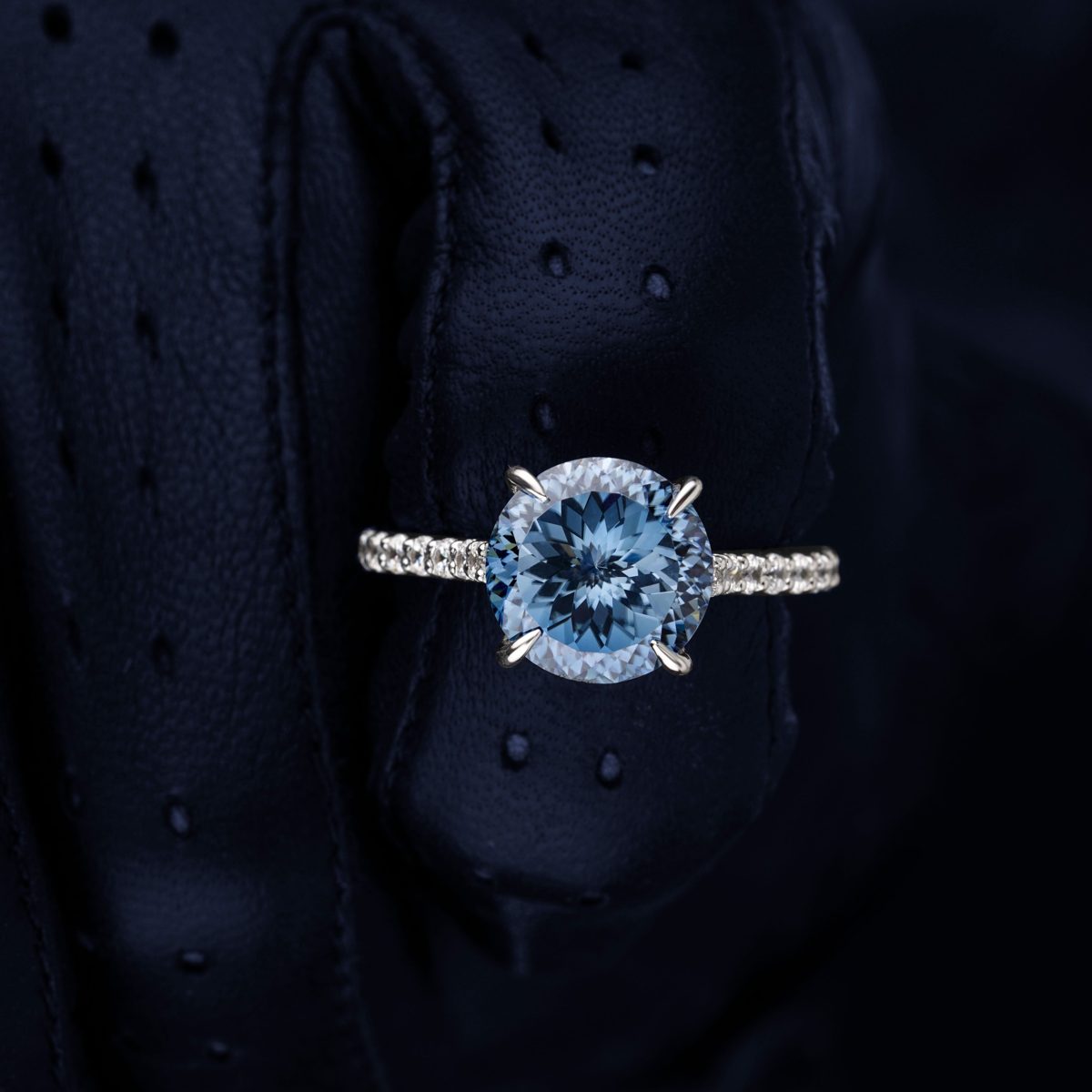 Round blue lab-grown diamond in a four-prong white gold setting with a pavé band, on dark navy leather