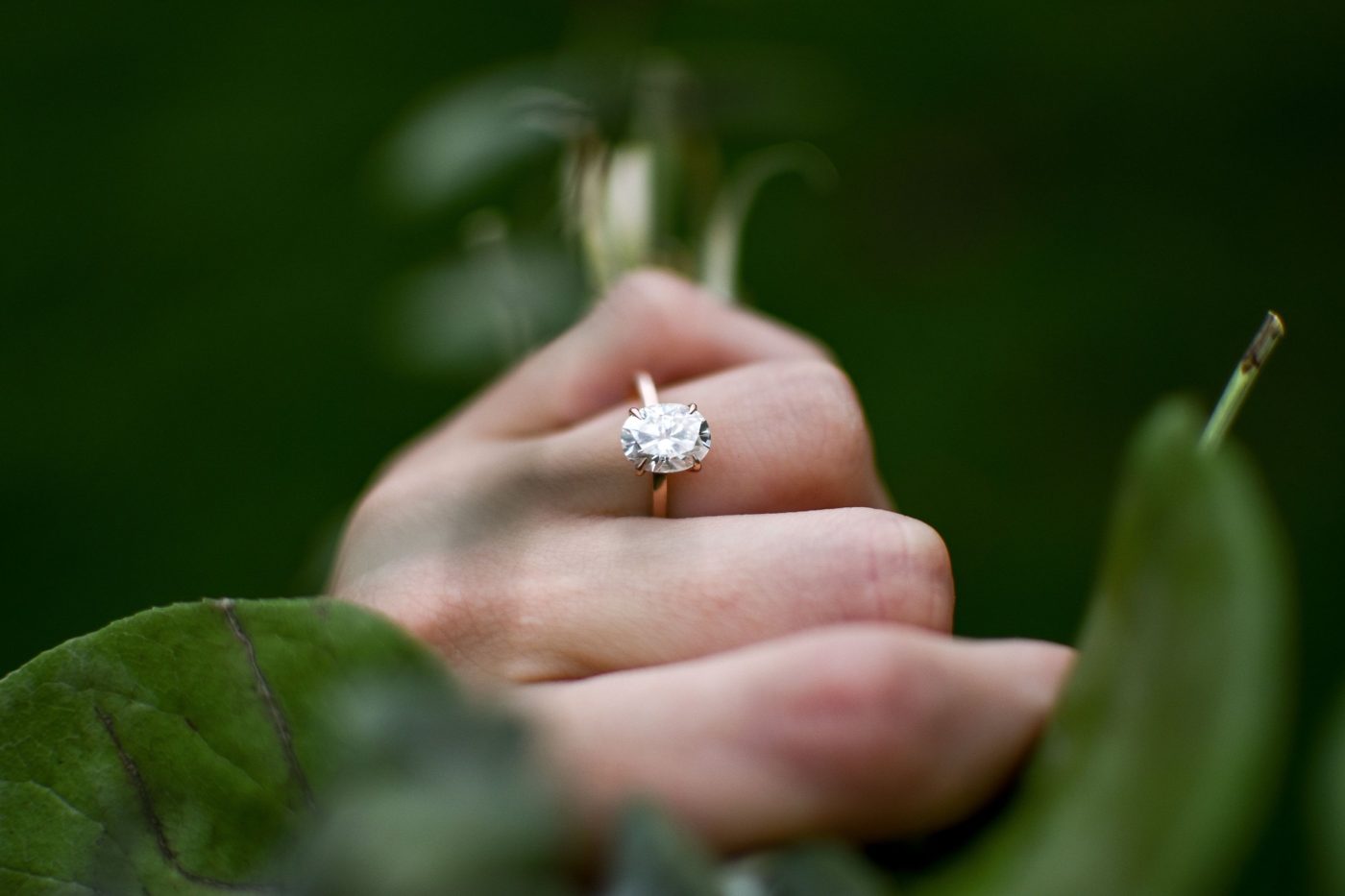 An oval solitaire in rose gold on a hand among green leaves